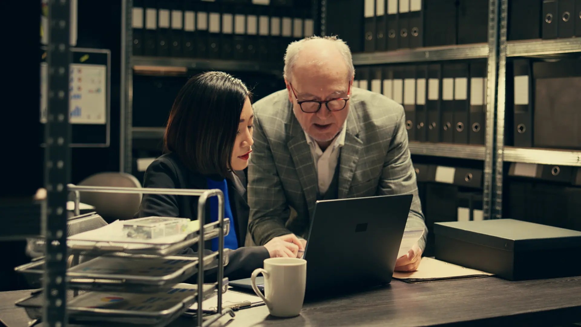 Two colleagues reviewing documents on a laptop in a records room lined with binders