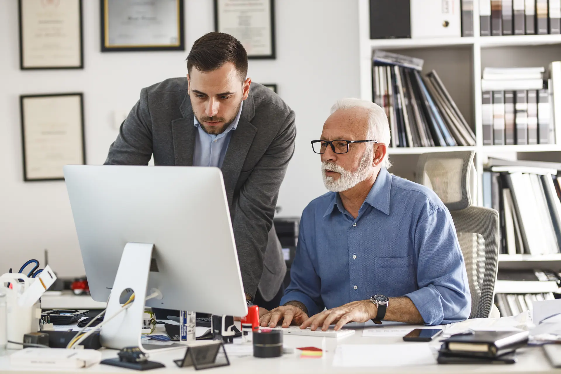 A younger colleague leaning in beside a senior professional at his desk, both reading the same screen